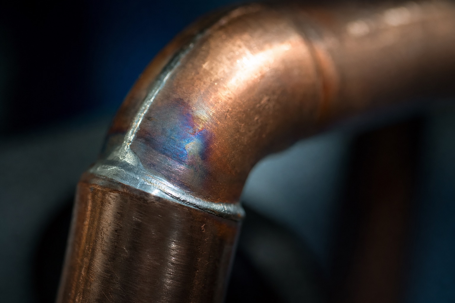 Macro photograph of a freshly soldered copper elbow joint, showing an even ring of silver solder around the seam.